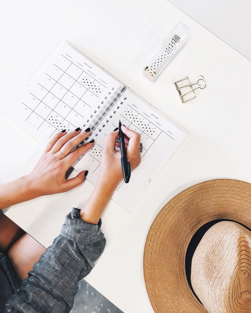 Woman writing in planner with wide-brim hat on desk.